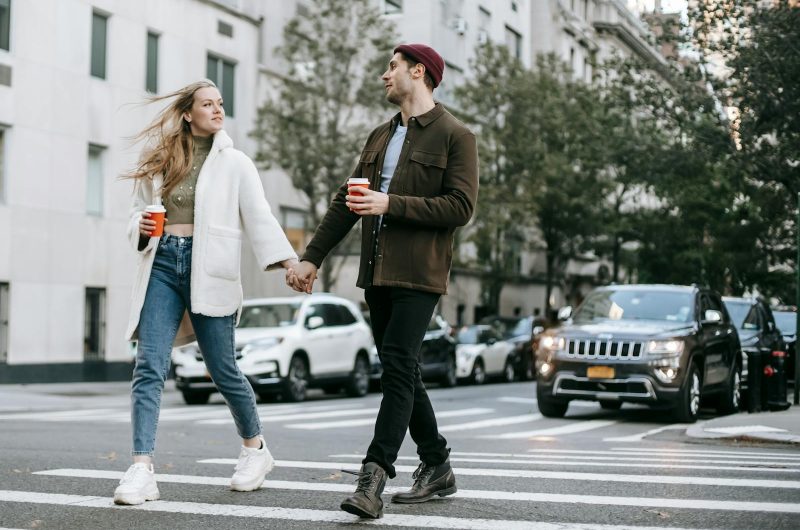 Young couple wearing warm jackets and with paper cups of hot drinks crossing road holding hands and looking at each other
