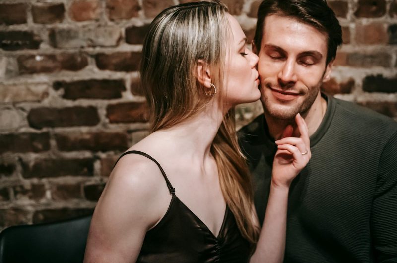 Young blond female kissing smiling boyfriend with closed eyes spending time together in cafe